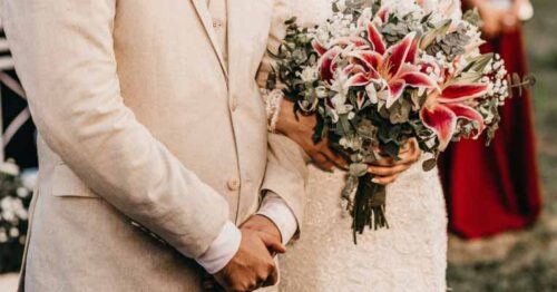 A lady dressed as a white lace bridal gown and holding a bouquet of flowers with prominent pink lilies.