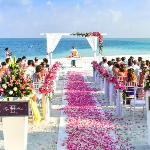 Beach wedding ceremony setup with white chairs and pink floral arrangements lining the aisle, ocean backdrop and a decorated archway.